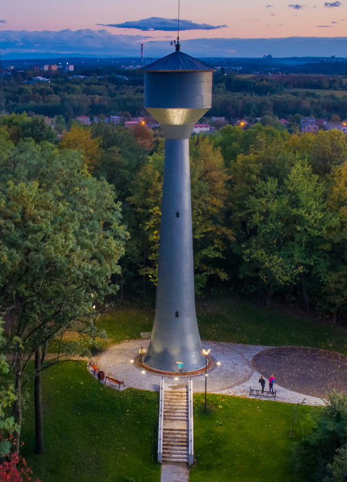 Nitowana wieża ciśnień w Będzinie-Grodźcu. Wieczorny widok z lotu ptaka na oświetlony teren wieży ciśnień. W centrum podświetlona nitowana, wysoka wieża w kształcie zadaszonego kielicha. Przed wieżą s 