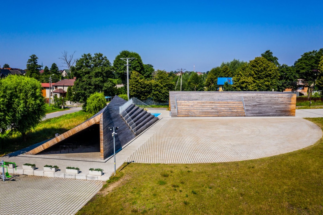 Widok z dalszej perspektywy na Centrum Aktywności Lokalnej w Kłokocinie – dwa drewniane wkomponowane w otoczenie budynki z siedziskami na zewnątrz na tle zieleni i błękitnego nieba. 