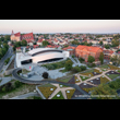 Hala Widowiskowo-Sportowa w Jaworznie. Zdjęcie zrobione w ujęciu z lotu ptaka. Z lewej strony podświetlony budynek hali i jego przedpole z alejkami i zielenią towarzyszącą. W tle widoczna panorama mia 