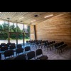 Wnętrze Centrum Aktywności Lokalnej w Kłokocinie – sala spotkań z ułożonymi konferencyjnie krzesłami – wykończone drewnem ściany i sufit, podłoga z dużych szarych kafelek, przeszklone wejście do obiektu. 