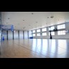 Sala sportowa od wewnątrz, podłoga wykończona drewnem, na jednej ścianie bramka do piłki, na drugiej ścianie dwa kosze do koszykówki, trzecia ściana z oknami, koszem do koszykówki i zegarem 