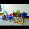Na zdjęciu znajduje się sala zabaw wraz z wyposażeniem. Ściany i podłogi są w jasnej kolorystyce. Siedziska dla dzieci i regały na zabawki są kolorowe. 