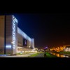 Widok hali Będzin Arena nocą w perpektywie liniowej. Po lewej oświetlony budynek hali z widocznym logo Będzina na telebimie. Po środku alejka nad bulwarami i rzeka. W tle oświetlony zamek na kolor fioletowy. Fotografia pochodzi ze zbiorów zgłaszającego 