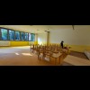 Sala przedszkolna z dużymi oknami, przez które wpada światło słoneczne. Widoczne stoliki i krzesełka dla dzieci. 