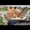 Zdjęcie z drona przedstawiające z góry drewniano-metalowe urządzania zabawowe Strefy, układające się w trzy połączone trójkąty, wokół zróżnicowana nawierzchnia, trawniki z drzewkami, ławki, leżaki. 