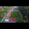 Widok z lotu ptaka. Stadion otoczony drzewami. W tle zabudowania. 