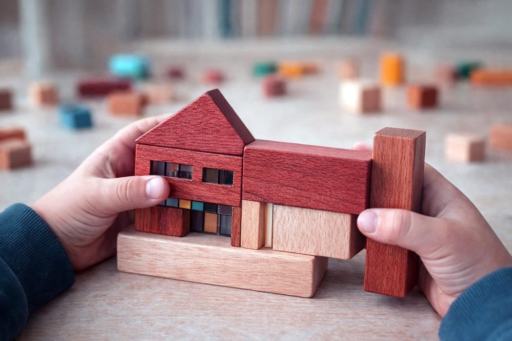 IDEA Dziecko trzyma drewniany model domu z kolorowymi detalami. Kompozycja pokazuje skalę wyobraźni dziecka i sposób postrzegania architektury przez najmłodszych.