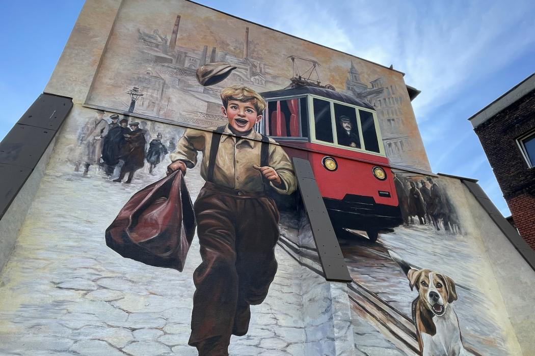 Mural na szczycie kamienicy: uśmiechnięty chłopiec z torbą biegnący brukowaną ulicą, obok pies, w tle czerwony tramwaj i sylwetka huty z kominami