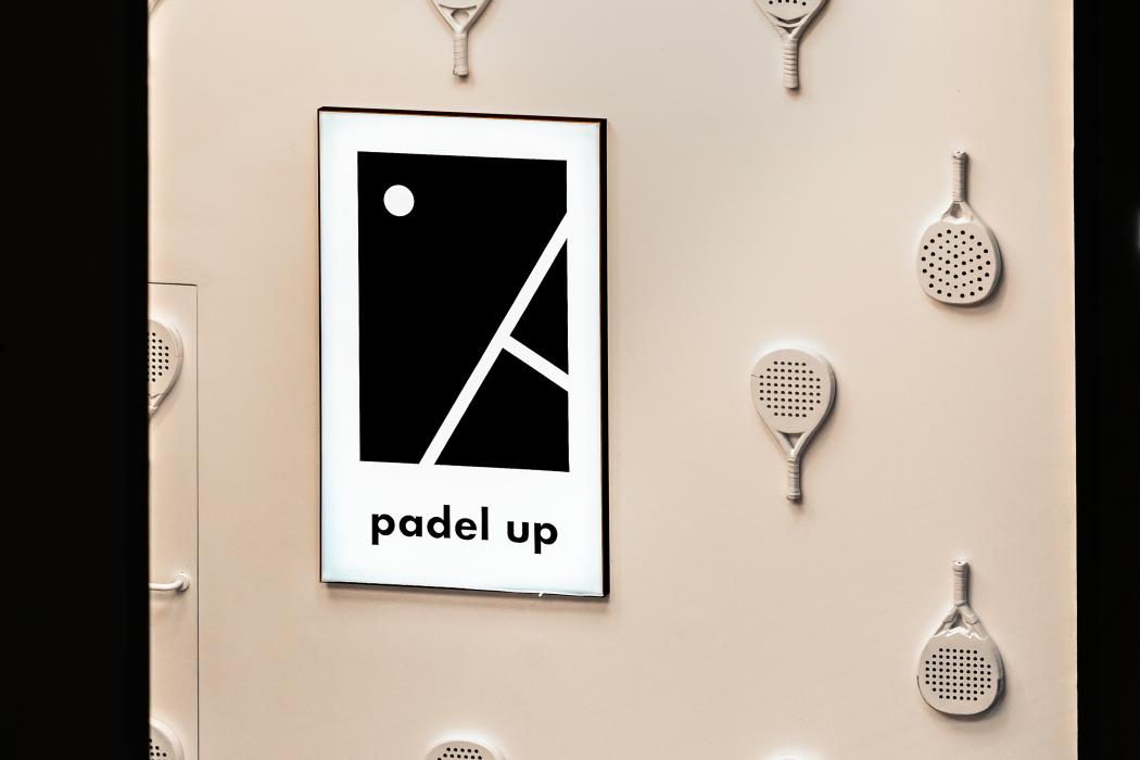 Widok strefy wejściowej. Biała ściana ozdobiona autorską instalacją z rakiet padlowych. Na środku czarno-biały plakat z logo klubu. U dołu widoczny fragment pomarańczowej pufy.