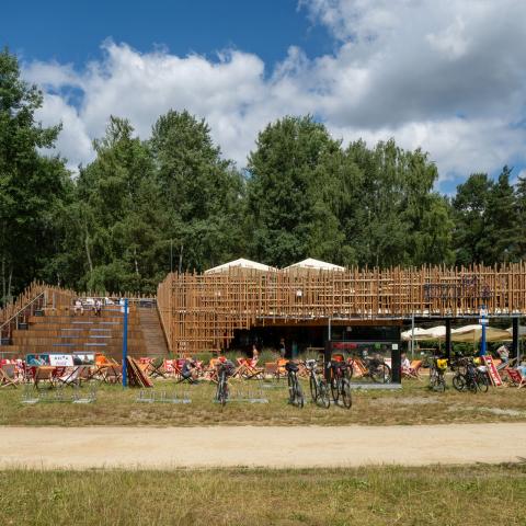 widok obiektu od przodu na tle lasu, drewniane elewacja i trybuna; w tle wysokie drzewa, na pierwszym planie ścieżka