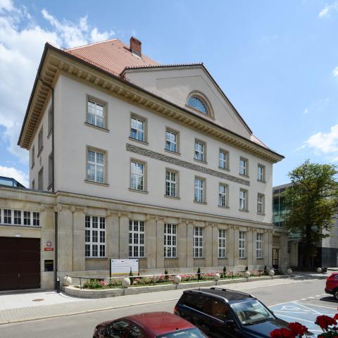 Widok w perspektywie ulicy Bankowej. Wyremontowana elewacja zabytkowego budynku. Dolna część z odnowionym detalem kamiennym, górna część wykończona tynkiem szlachetnym w kolorze piaskowym. Elewacja zwieńczona kamiennym gzymsem , ponad gzymsem trójkątny naczółek. Budynek kryty dachówką o naturalnej, ceglastej barwie.