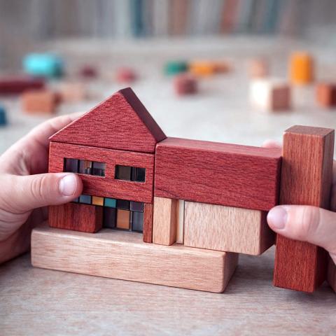 IDEA Dziecko trzyma drewniany model domu z kolorowymi detalami. Kompozycja pokazuje skalę wyobraźni dziecka i sposób postrzegania architektury przez najmłodszych.