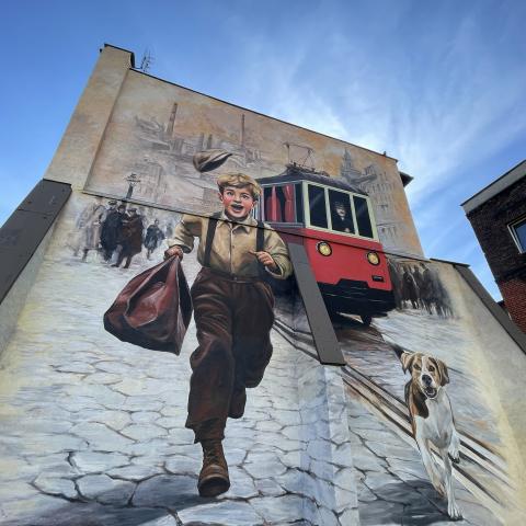 Mural na szczycie kamienicy: uśmiechnięty chłopiec z torbą biegnący brukowaną ulicą, obok pies, w tle czerwony tramwaj i sylwetka huty z kominami