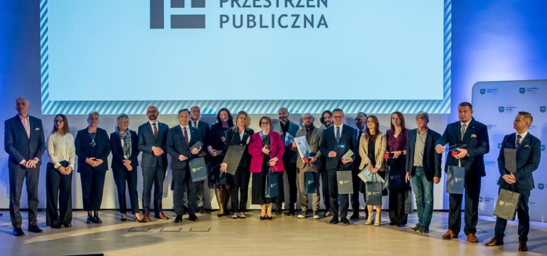Zdjęcie grupowe laureatów konkursu na najlepszą przestrzeń publiczna województwa śląskiego, na tle slajdu z logiem konkursu. 