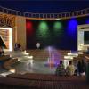 Wnętrze atrium tężni solankowej po zmierzchu z centralnie zlokalizowaną fontanną posadzkową z iluminacją świetlną oraz amfiteatralną widownią wypełnioną kuracjuszami. W tle oświetlone iluminacyjnie ściany tężni wypełnione tarniną, po której spływa solanka 