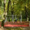 Widok na czerwony plac z urządzeniami do streetworkoutu na którym bawi się trójka dzieci. W centrum drabinki i urządzenia do ćwiczeń. Plac otoczony drzewami. Na pierwszym planie czerwona ścieżka.