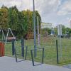 Zdjęcie przedstawia ogrodzony plac zabaw na świeżym powietrzu. Na pierwszym planie znajduje się wejście na teren placu, zabezpieczone metalową furtką oraz chodnik z kostki brukowej. Po lewej stronie stoi nowoczesny kosz na śmieci w obudowie z drewnianych listew oraz huśtawka z metalową ramą i drewnianym siedziskiem. W centrum placu widać czerwoną siatkę wspinaczkową w kształcie stożka. Po prawej stronie chłopiec zjeżdża ze zjeżdżalni. W tle rosną wysokie drzewa iglaste, a za nimi znajdują się niskie budynki