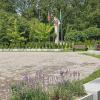 Widok na pomnik w parku otoczonym zielenią i flagami. Na pierwszym planie rabaty z kwitnącą lawendą w betonowych donicach, za nimi brukowany plac. W tle widać monument z uniesionymi dłońmi oraz flagami: Polski, Unii Europejskiej i Ukrainy. Otoczenie pełne niskiej roślinności, drzew oraz ławek, a wszystko wkomponowane w naturalny leśny krajobraz.