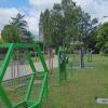 Zdjęcie przedstawia fragment parku z zamontowanymi zielonymi konstrukcjami sensorycznymi, służącymi do zabawy i rozwijania zmysłów, szczególnie dla dzieci. Na pierwszym planie widać dużą zieloną ramę w kształcie sześciokąta z zawieszonymi na prostych drutach metalowymi kulkami. Dalej widać kolejne podobne konstrukcje – ramy z elementami do dotykania, przesuwania i obserwacji. Po prawej stronie znajduje się trawnik, a w tle drzewa i alejka prowadząca przez park. W oddali widoczna jest wieża kościoła. 