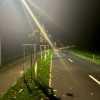 Oświetlona nocą velostrada i ciąg pieszy, rozdzielone pasem zieleni na którym rosną młode drzewa zabezpieczone drewnianymi podporami. Trasa biegnie łagodnym łukiem przez teren zielony. Na asfaltowej nawierzchni widoczne są białe linie rozdzielające pasy ruchu oraz leżące pojedyncze jesienne liście. Jasne światło lamp ulicznych rozświetla trasę, zapewniając bezpieczeństwo i komfort rowerzystom po zmroku.