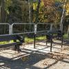 Dwie ławki oraz stojaki rowerowe ustawione na utwardzonej nawierzchni przy velostradzie, w otoczeniu jesiennych drzew. Obok znajduje się kosz na śmieci oraz samoobsługowa stacja naprawy rowerów. Całość oddzielona jest metalową barierką od zalesionego terenu. Elementy te stanowią przykład małej architektury towarzyszącej infrastrukturze velostrady, zapewniając miejsce odpoczynku i wsparcie dla rowerzystów. Zdjęcie wykonano w słoneczny dzień.