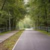 Zdjęcie przedstawia asfaltową velostradę po prawej stronie oraz węższy ciąg pieszy po lewej, oddzielone pasem zieleni. Obie ścieżki biegną równolegle przez zalesiony teren, otoczone są metalowymi barierkami. Wzdłuż ciągu pieszego stoją latarnie. Droga prowadzi w głąb lasu, tworząc przyjazną trasę rekreacyjną.