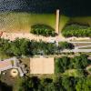 Od góry widać czarną toń jeziora, przechodzącą w piaszczystą plażę, następnie ścieżka w zieleni. Na plaży leżaki i opalający się ludzie. Centralnie drewniana wieża ratowników