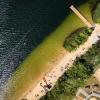 Widok z góry fragmentu plaży, biegnącej po skosie kadru. Od lewej widać jezioro, Pas piaszczystej plaży z pomostem, zielone korony dużych drzew, pochylnię dla niepełnosprawnych, W dole boisko do siatkówki plażowej oraz altany na łące.