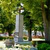 Zdjęcie przedstawia pomnik w centralnej części parku, otoczony zielenią i kwiatami. Monument ma formę prostokątnego, pionowego obelisku. Na szczycie pomnika umieszona jest figura orła z rozpostartymi skrzydłami. U podnóża pomnika widoczny jest wieniec oraz elementy dekoracyjne. Całość otaczają drzewa liściaste, rabaty z kwiatami oraz niska roślinność ozdobna. W tle widoczne elementy małej architektury.