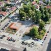 Widok na  centralny plac miejscowości – Rynek w Koniecpolu. Na pierwszym planie widoczny jest wybrukowany plac z wyznaczonymi miejscami parkingowymi oraz niewielkim pawilonem. W centrum kadru rozciąga się teren zielony z licznymi drzewami, alejkami spacerowymi i ławkami. Otaczające ulice tworzą prostokątny obrys placu. Dzień jest słoneczny, widoczne są cienie rzucane przez drzewa i budynki.