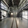 Na zdjęciu: wnętrze budynku Warsztatu Elektrycznego - sala główna. Posadzka szlifowana w kolorze szarym z szynami w części centralnej. Na ścianie po lewej stronie przeszklenie na wysokości około 2m, po prawej antresola. Konstrukcja antresoli stalowa w kolorze grafitowym, ściany z widoczna konstrukcją szachulcową w kolorze grafitowym. Pozostała część ścian malowana na biało.