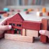 IDEA Dziecko trzyma drewniany model domu z kolorowymi detalami. Kompozycja pokazuje skalę wyobraźni dziecka i sposób postrzegania architektury przez najmłodszych.