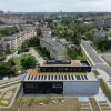 Widok z lotu ptaka od zachodu. Widoczny parking, ogrodzony teren reakreacyjny oraz budynek. Widoczna instlacaja paneli fotowoltaicznych oraz dach zielony.