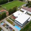 Zdjęcie lotnicze przedstawiające kompleks szkolno-sportowy położony wśród terenów zielonych. W centralnej części znajduje się nowoczesny budynek z jasnym dachem, połączony ze starszym budynkiem o czerwonym dachu. Obok widoczne są pełnowymiarowe boisko piłkarskie z zieloną murawą oraz wielofunkcyjne boisko sportowe. Przed budynkiem znajduje się parking z zaparkowanymi samochodami, w tym wyznaczone miejsca dla osób z niepełnosprawnościami. Całość otaczają drzewa i niska zabudowa.