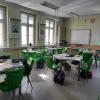 Sala lekcyjna z zielonymi krzesłami i stolikami ustawionymi w grupach. Na wyposażeniu znajdują się tablica interaktywna, projektor oraz materiały edukacyjne na stołach.