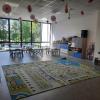 Sala przedszkolna ze stolikami i krzesełkami ustawionymi w grupy. Na wyposażeniu znajdują się meble z zamykanymi szafkami oraz zabawki i książki, na środku sali znajduje się dywan przedstawiający miasto z drogami, budynkami i zielenią. Centralna ściana sali jest w całości przeszklona z widokiem na parking i zieleń otaczającą budynek.