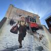 Mural na szczycie kamienicy przedstawiający uśmiechniętego chłopca z torbą biegnącego brukowaną ulicą, obok średniej wielkości pies, w tle czerwony tramwaj i sylwetka huty z kominami