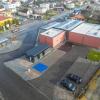 Widok centrum przesiadkowego od parkingu z drona. Na zdjęciu widoczne jest centrum Jejkowic wraz z centrum przesiadkowym. Przez środek przebiega droga powiatowa. Na środku widoczna asfaltowa zatoka autobusowa, wysepka z wiatą przystankową. Z prawej góry widoczne zabudowania centrum przesiadkowego. Widoczne są białe dachy, elewacja imituje czerwoną cegłę. Z prawego dołu widoczny jest parking dla samochodów. Wjazd na parking prowadzi z drogi wewnętrznej.