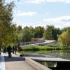Widok na marinę żeglarską z promenady przy marinie kajakowej. Wznosząca się drewniana promenada w zieleni.