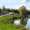 Widok na wznosząca się drewnianą promenadę wkomponowaną w zieleń, po prawej tafla jeziora i zacumowane łodzie.
