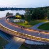 Widok na promenadę ze spacerowiczami z góry w wieczornym oświetleniu. Na pierwszym planie prześwit pomiędzy częścią szkoleniową a wiatami na łodzie. W tle jezioro, w oddali sylweta miasta.