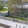 Widok na budynek od strony parkingu. Na pierwszym planie widać biały, parterowy budynek techniczny. Za nim znajduje się zielone patio, z szeregiem wysokich drzew.