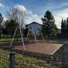 Plac zabaw z huśtawką ustawioną na gumowej nawierzchni amortyzującej. W tle widoczne są drzewa, dom jednorodzinny oraz ogrodzenie.