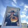 Mural na szczycie budynku przedstawiający mężczyznę w mundurze grającego na trąbce, w tle huta z kominami i tłum ludzi, kompozycja w łukowym obramieniu