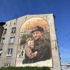 Mural na elewacji kamienicy: hutnik w czapce trzymający małe dziecko, oboje uśmiechnięci, w tle kominy huty i przedwojenna ulica, kompozycja w łukowym obramieniu