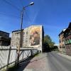 Mural z hutnikiem z dzieckiem rękach na elewacji kamienicy przy ul. Szpitalnej 23, widok od strony ulicy z infrastrukturą hutniczą po lewej stronie oraz jezdnią i fragmentem starej kamienicy po prawej