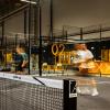Zbliżenie na siatkę z logo "padel up". W rozmytym tle na przeszklonym korcie grają dwaj mężczyźni. W głębi żółtopomarańczowa lada.