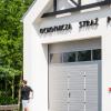 Fragment budynku z jasnymi ścianami i charakterystyczną konstrukcją dachową z ciemnymi belkami drewnianymi. Na fasadzie widoczny napis „OCHOTNICZA STRAŻ POŻARNA". Budynek posiada duże bramy garażowe w kolorze szarym z przeszkleniami w górnej części. Przed budynkiem przebiega osoba w ciemnej koszulce i jasnych spodniach. W tle widoczna jest zieleń drzew, a całość prezentuje się na tle błękitnego nieba.